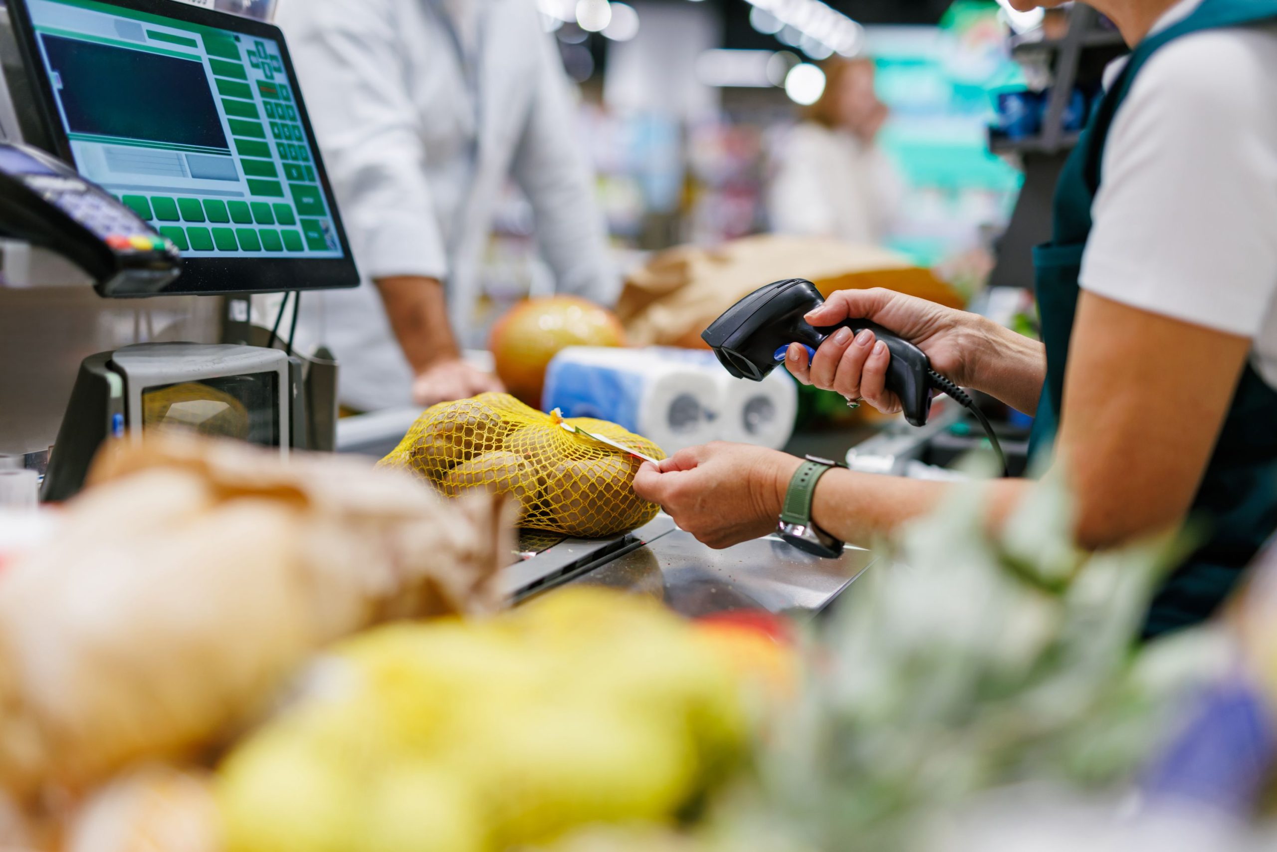 Supermarket cashier scanning potatoes at the checkout, while a customer waits in the background.